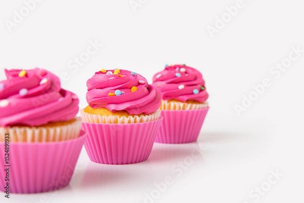 Fototapeta Three pink frosted cupcakes with colorful sprinkles on white background
