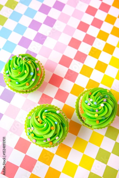 Fototapeta Green frosted cupcakes with colorful sprinkles on checkerboard background, top view