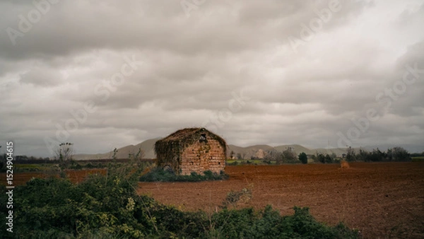 Fototapeta An abandoned stone house in the middle of a plowed field, under a dramatic cloudy sky. In the background you can see soft hills and country vegetation. Silent and melancholic atmosphere.