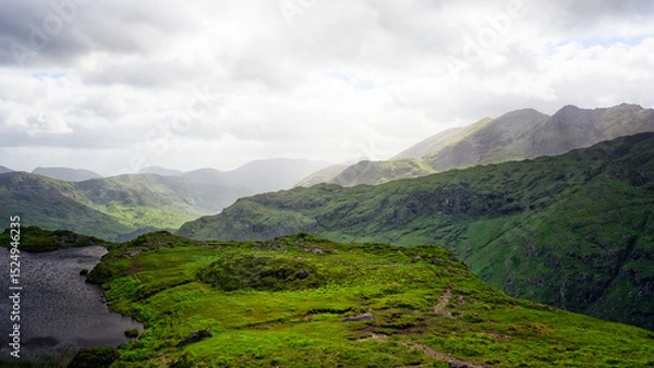 Fototapeta A scenic vista shows layers of rolling green hills and a tranquil lake from an overlook in the Gap of Dunloe, County Kerry. Overcast sky with scattered light accentuates the landscape.