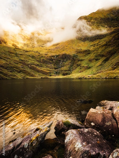 Fototapeta This showcases the still waters of Lough Callee, reflecting the surrounding hills. A small waterfall cascades down the slope into the calm lake, nestled in the Irish countryside.