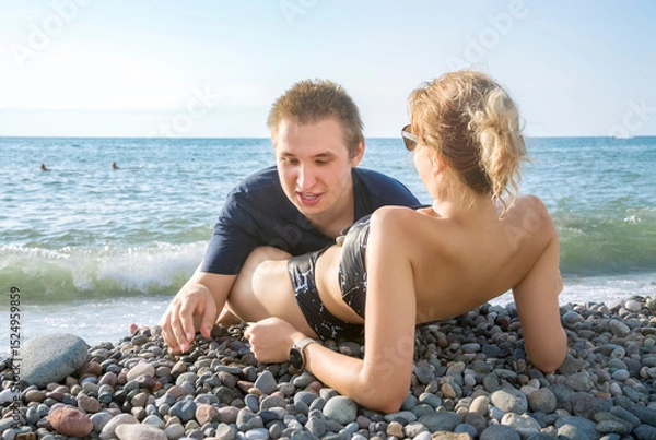 Fototapeta A young man and a girl are talking, lying on a pebble beach at the water's edge