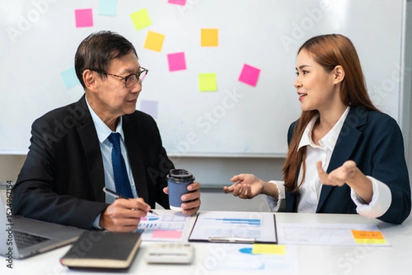 Obraz Two people in suits are talking at a desk with a white board behind them