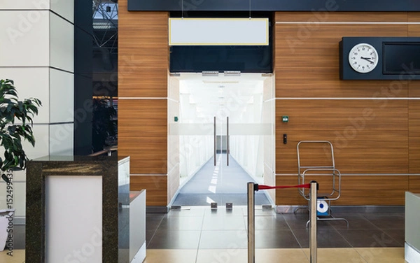 Obraz Glass doors open to empty jet bridge inside bright airport terminal. Clean floors, wooden wall panels with clock and flight information display, organized airport space for boarding passengers