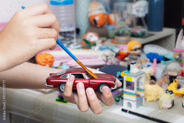 Fototapeta Hand cleaning dust off a red toy car with small brush on cluttered tabletop. Concept of dusting, toy maintenance, cleaning miniature models, detailed care, indoor hobby activity