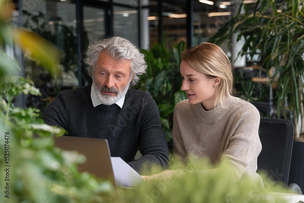 Obraz Older man and younger woman collaborate on laptop, seated at table amidst indoor plants.  Image depicts teamwork and modern office environment