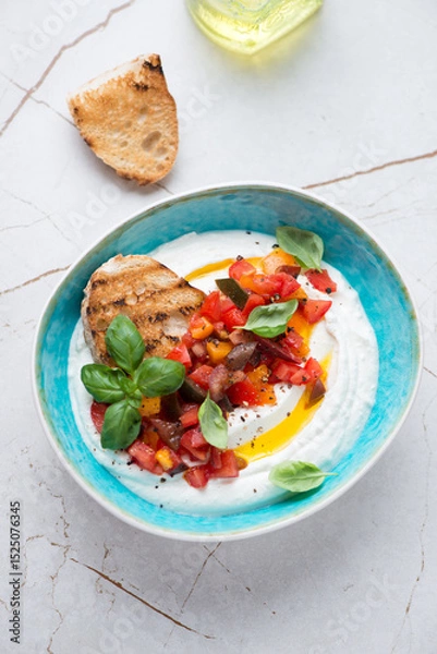 Fototapeta Turquoise bowl with ricotta dip, chopped tomatoes and ciabatta, vertical shot on a white stone background, elevated view