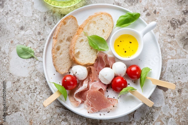 Fototapeta White plate with ciabatta, prosciutto and caprese skewers, horizontal shot on a light-brown granite background, elevated view
