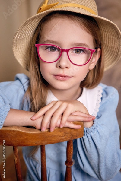 Fototapeta A charming young girl of European appearance in a blue dress with a white lace collar sits with her hands on the back of a chair. The child is wearing a straw hat and glasses with bright red frames.