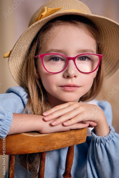 Fototapeta An attractive young girl of Slavic appearance with light-brown hair, wearing glasses with pink frames and a straw hat with a yellow ribbon, sits with her arms crossed on the back of a chair 