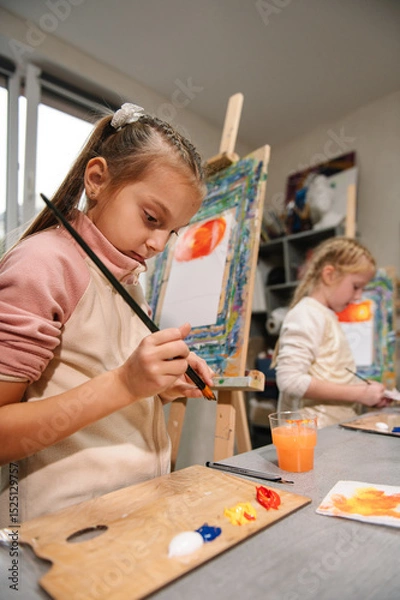 Fototapeta Education and learning concept. School kids drawing with brush landscapes on easel. Girls enjoy painting in a workshop. Art lesson. Mix and combine colors. Children drew a picture in art class.