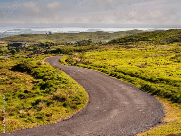 Fototapeta Small s shaped road with green fields on a side and lake with many small islands under cloudy sky in the background. Stunning nature scenery in Connemara, Ireland. Travel and tourism.