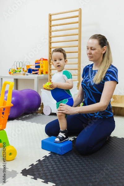 Fototapeta Physiotherapist assists a young boy patient wearing in special shoes in maintaining balance during a physical rehabilitation activity with fitness block