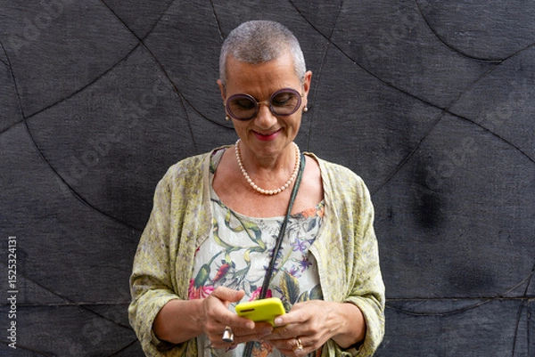 Obraz elegant smiling mature woman with shaved grey hair texting on cell phone in front of black wall