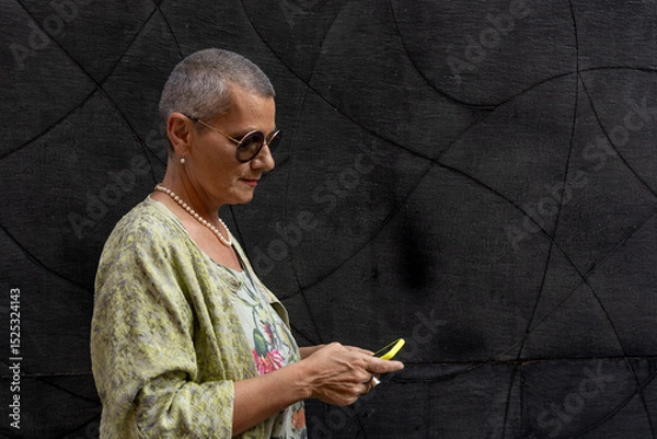Obraz elegant smiling mature woman with shaved grey hair texting on cell phone in front of black wall