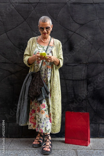 Obraz elegant smiling mature woman with shaved grey hair texting on cell phone in front of black wall