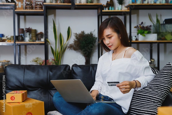 Obraz Asian woman holding a credit card while shopping online with a laptop at home. Concept of e-commerce, lifestyle, and digital payment in a cozy modern living space.
