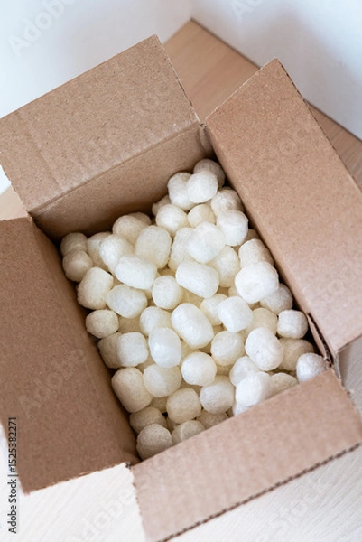Fototapeta Open cardboard box filled with eco-friendly corn-based packing peanuts on a light wooden surface. Sustainable packaging for safe and conscious shipping, promoting zero-waste