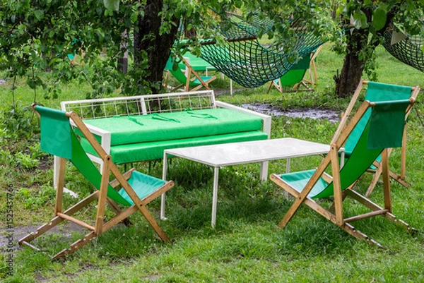 Fototapeta Folding wooden chairs, a summer sofa and a table on a lawn in the garden. Recreation area in park.