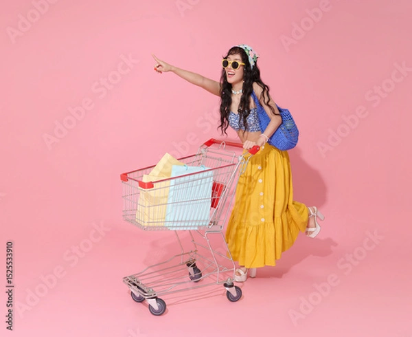 Fototapeta Happy smiling young asian woman dressed in summer clothes pushing trolley cart and holding shopping bag summer sale isolated on pink background.