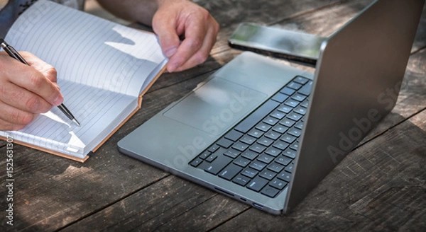 Fototapeta Businessman or freelancer writing in notebook while working outdoors on laptop at wooden table. Creative remote work or planning concept