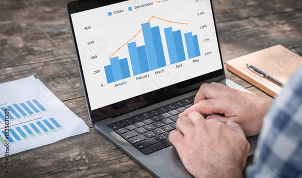 Fototapeta Male entrepreneur analyzing monthly conversion and click-through analytics on laptop at wooden table workspace. Remote work and data analysis concept