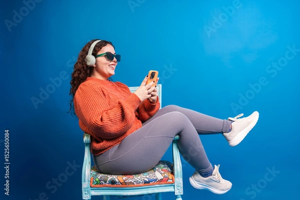 Fototapeta Plus size woman using mobile phone while listening music with headphones and wearing sunglasses, sitting on a chair