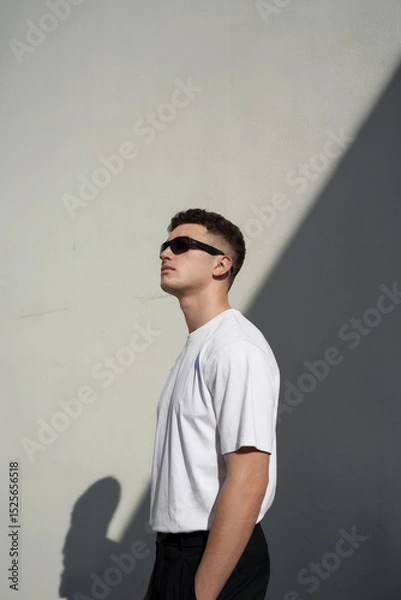 Fototapeta A minimalist portrait photograph of a young man in profile view, looking upward against a plain light gray wall. He wears a plain white crew neck t-shirt and black sunglasses. 