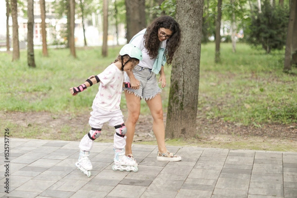 Fototapeta Mother teaching her daughter how to rollerblade in a park. The girl wears protective gear and helmet. A joyful and bonding outdoor moment filled with love and support.