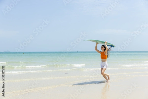 Fototapeta Joyful young woman walks out of water at sunny beach, holding surfboard. She wears bright orange top and white shorts, with clear blue sky in background.