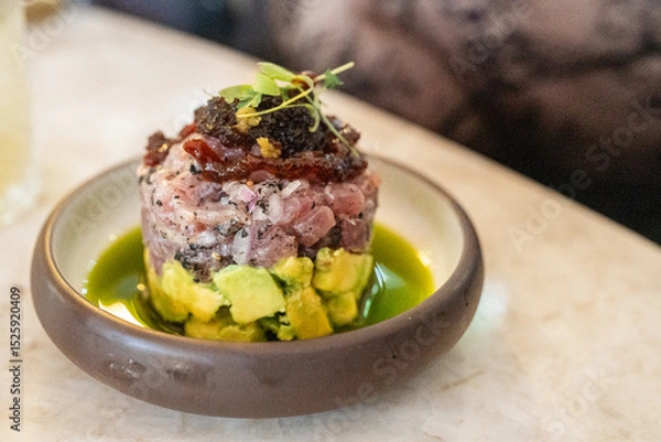 Obraz A beautifully plated tartare featuring fresh fish layered with creamy avocado. The dish is garnished with toppings and presented in a stylish bowl, showcasing vibrant colors and textures.