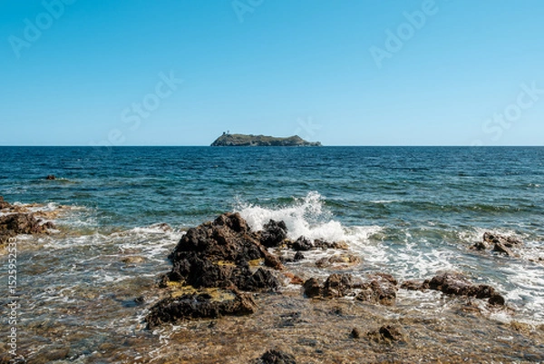 Fototapeta Waves splashing onto the rocky coastline of the northern tip of Cap Corse on the Mediterranean island of  Corsica with the island of Giraglia in the distance