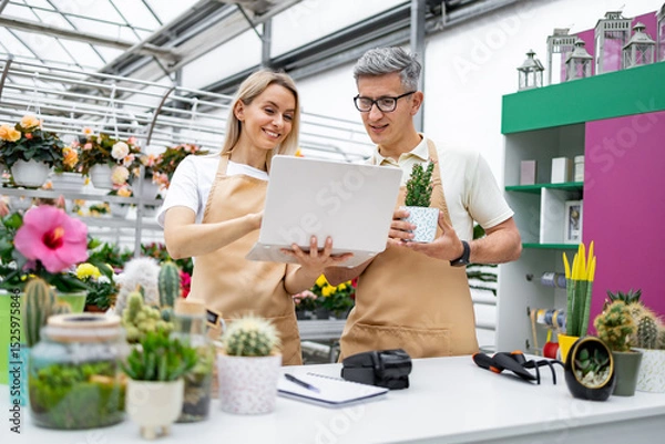 Obraz Two florists, a man and a woman, look at a laptop in a plant shop, discussing a small plant in a pot at the table.