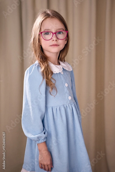 Fototapeta Backlit portrait of a young girl with long curly hair wearing glasses in a pink frame. She is wearing a blue dress decorated with buttons and a white lace collar. In the background is a light curtain.