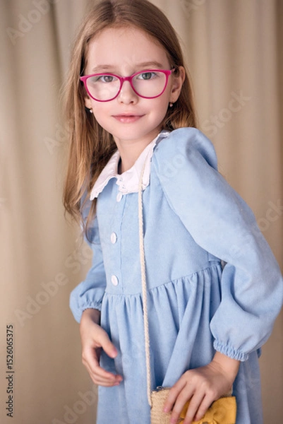 Fototapeta A beautiful young girl in a blue dress with a white collar and glasses coquettishly adjusts a small straw bag with her hand. She has long blond hair and looks at the camera with a slight smile. 