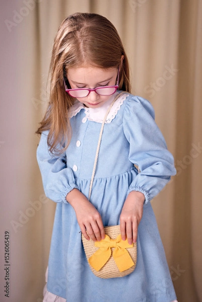 Fototapeta A young girl with long blonde hair is looking for something in her small straw bag with a yellow bow. The atmosphere of the photo conveys a sense of childish naivety, creating a warm mood.