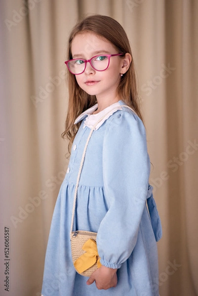 Fototapeta A charming little girl with a cute wicker handbag over her shoulder stands half-turned on a milky fabric background. The child in glasses is dressed in an elegant velvet dress with a white collar
