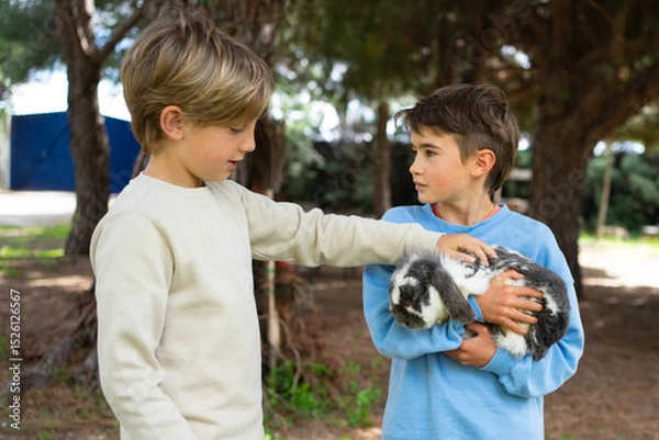 Fototapeta Children petting rabbit in a forest: two young boys showing affection to a cute bunny