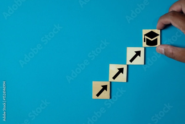 Obraz Hand arranging wooden blocks in steps with arrows leading to a graduation cap icon on blue background.Concept for educational goals,skill improvement, learning progress,personal development.