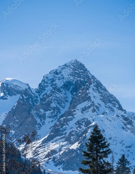 Fototapeta Majestic snow-capped mountain peak stands tall against clear blue sky, with rugged terrain and patches of snow covering its slopes. Karachay-Cherkessia. Dombay 2025. Nature concept for design.
