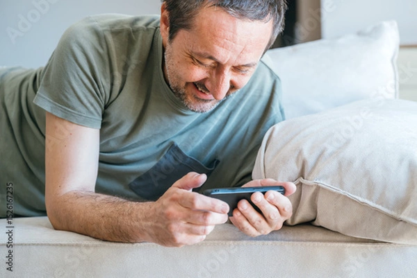 Fototapeta Casual male watching entertaining social media content on mobile phone device while laying on living room sofa