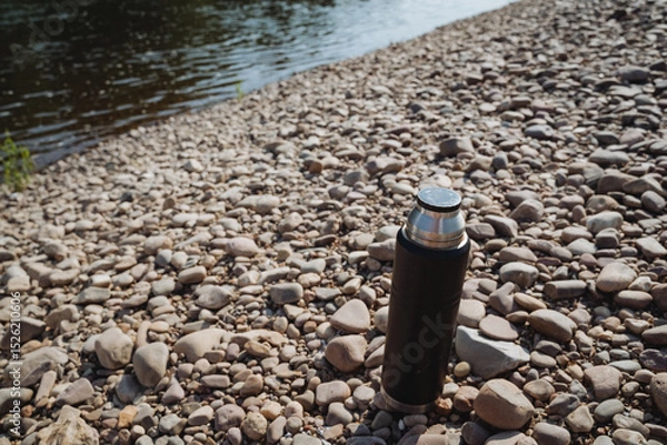 Obraz A Stainless Steel Thermos Bottle rests gracefully on the Pebbly Riverbank, inviting you to embark on an adventure filled with outdoor activities like hiking, camping, and serene picnics by the beach