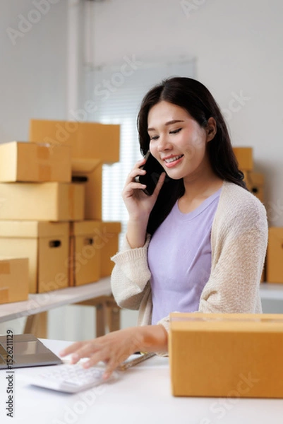 Fototapeta Asian woman entrepreneur managing retail business from home office talking on phone and calculating costs