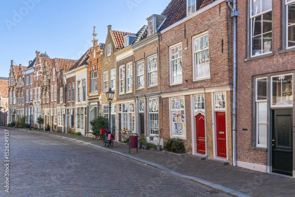 Obraz This captivating photo showcases a charming, historic street in Dordrecht, Netherlands, lined with classic Dutch gabled houses on a sunlit cobblestone path. Dordrecht, Netherlands. 16 March 2025.