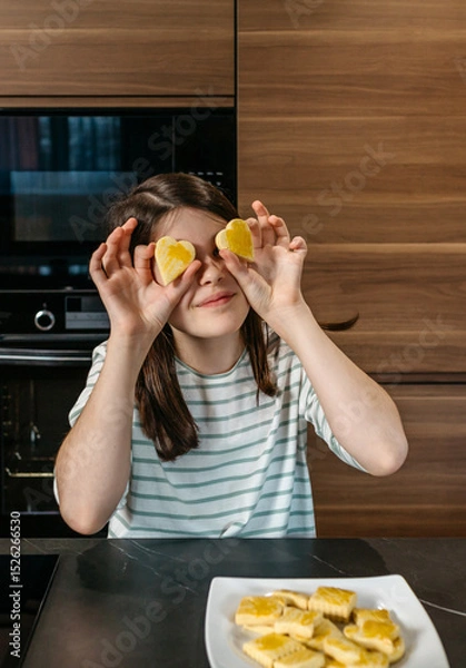 Fototapeta Young girl having fun in the kitchen, holding two heart shaped cookies over her eyes, expressing playfulness and enjoyment. Happy little girl looking at camera while covering her eyes with biscuits.