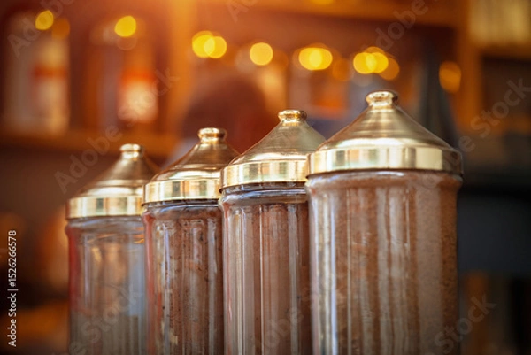 Fototapeta Clear glass dispensers with ornate gold tops stand in a warm, dimly lit shop, showcasing Turkish coffee. The focus is on the intricate details of these traditional containers.