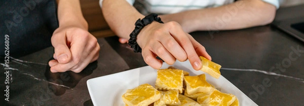 Fototapeta Banner of unrecognizable girl is picking a freshly baked cookie from white plate on kitchen countertop next to another child hands waiting to eat