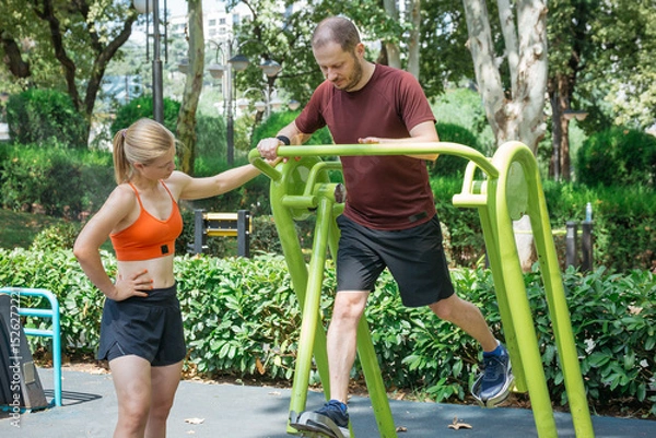 Obraz Man exercising on outdoor fitness equipment in a park, assisted by a female trainer.  They are using a leg press machine for a lower body workout. Healthy lifestyle and fitness concept.