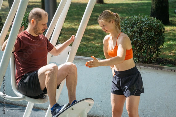 Obraz A young female fitness instructor assists a man during a leg workout on outdoor gym equipment. They are exercising in a sunny park, showcasing a healthy active lifestyle.