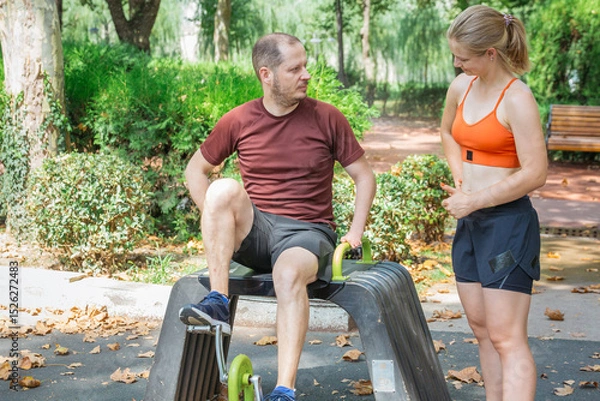 Obraz Man exercising leg press machine outdoors in a park. Woman assists or instructs him.  Focus on health, fitness, and teamwork.